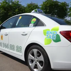 Biała taksówka EKO TAXI z zielonym logo i numerem telefonu na drzwiach.