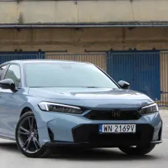 Nowy Honda Civic Sport w kolorze szarym, z czarnymi felgami, zaparkowany przed industrialnym budynkiem z niebieskimi bramami.