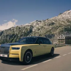 Żółty Rolls-Royce Phantom przemierza górską drogę. W tle majestatyczne szczyty i zabytkowy hotel.