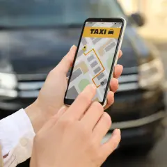 Dłoń trzyma smartfon z aplikacją taxi i mapą, na tle samochodu.