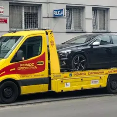 Żółta laweta z napisem "Pomoc drogowa" przewozi czarny samochód przed budynkiem policji. Konfiskata samochodu w akcji.