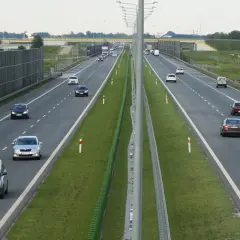Samochody poruszają się po wielopasmowej drodze ekspresowej. Po lewej stronie widać wysokie ekrany akustyczne, a po prawej zielone pasy zieleni i znaki drogowe.
