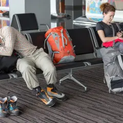 Zmęczony turysta śpi na lotnisku, obok niego plecak. Czy to efekt overbookingu? Kobieta obok sprawdza telefon.
