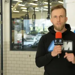 Mężczyzna z mikrofonem w serwisie BMW, informuje o akcji serwisowej. W tle widać samochody i podnośniki.