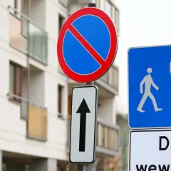 Znak zakaz postoju nad strzałką kierunkową, obok niebieskiej tablicy z piktogramami pieszych i samochodu. Budynek mieszkalny w tle.