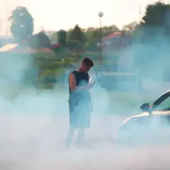 Młody mężczyzna z telefonem w dłoni stoi w kłębach dymu z palonych opon. Samochód obok, jakby po awarii uszczelniaczy zaworowych, generuje dym.