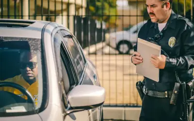 Policjant wręcza kierowcy mandat za kolizję. Mężczyzna w okularach przeciwsłonecznych siedzi w samochodzie.