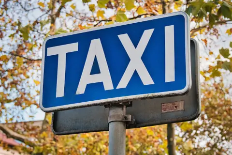 Niebieski znak TAXI z białymi literami na tle jesiennych drzew.