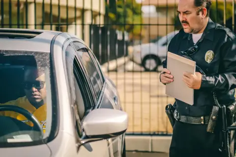 Policjant wręcza kierowcy mandat za kolizję. Mężczyzna w okularach przeciwsłonecznych siedzi w samochodzie.