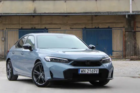 Nowy Honda Civic Sport w kolorze szarym, z czarnymi felgami, zaparkowany przed industrialnym budynkiem z niebieskimi bramami.