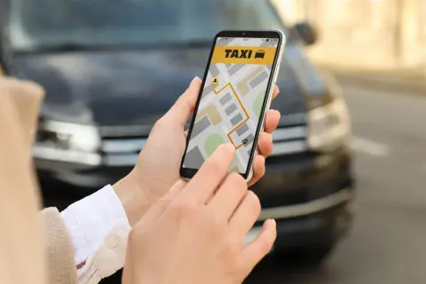 Dłoń trzyma smartfon z aplikacją taxi i mapą, na tle samochodu.
