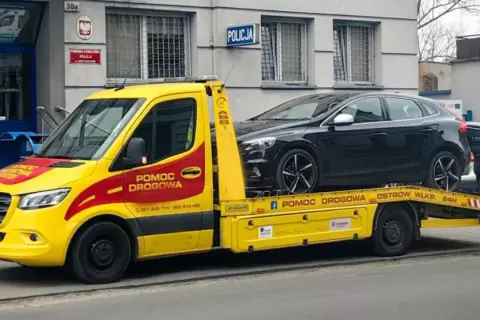 Żółta laweta z napisem "Pomoc drogowa" przewozi czarny samochód przed budynkiem policji. Konfiskata samochodu w akcji.
