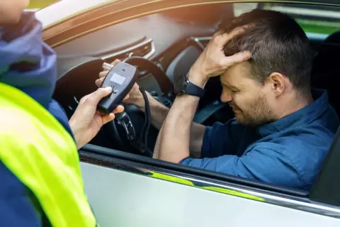 Policjant mierzy kierowcy ile promili. Mężczyzna zmartwiony trzyma się za głowę.