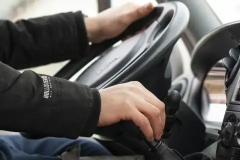 Ręce kierowcy na kierownicy i drążku zmiany biegów. Na drodze obowiązuje zakaz wyprzedzania.