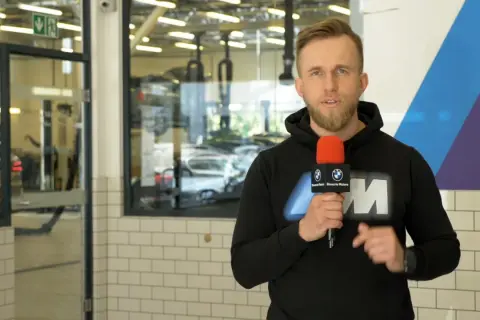 Mężczyzna z mikrofonem w serwisie BMW, informuje o akcji serwisowej. W tle widać samochody i podnośniki.