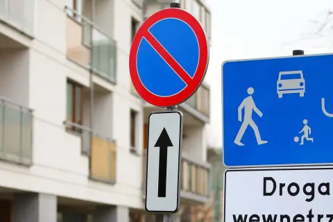 Znak zakaz postoju nad strzałką kierunkową, obok niebieskiej tablicy z piktogramami pieszych i samochodu. Budynek mieszkalny w tle.