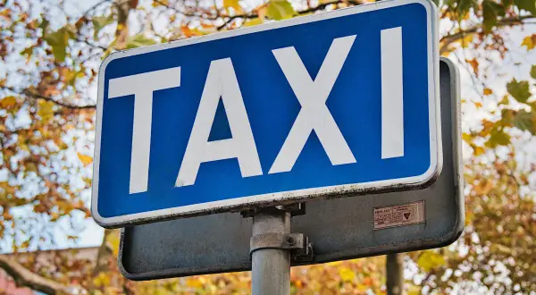Niebieski znak TAXI z białymi literami na tle jesiennych drzew.