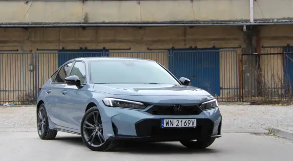 Nowy Honda Civic Sport w kolorze szarym, z czarnymi felgami, zaparkowany przed industrialnym budynkiem z niebieskimi bramami.