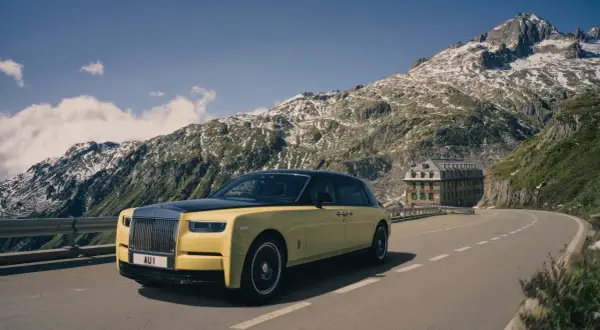 Żółty Rolls-Royce Phantom przemierza górską drogę. W tle majestatyczne szczyty i zabytkowy hotel.