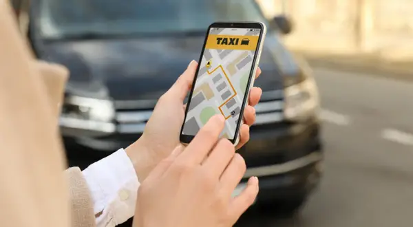Dłoń trzyma smartfon z aplikacją taxi i mapą, na tle samochodu.
