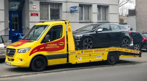 Żółta laweta z napisem "Pomoc drogowa" przewozi czarny samochód przed budynkiem policji. Konfiskata samochodu w akcji.