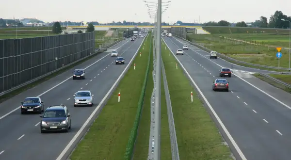 Samochody poruszają się po wielopasmowej drodze ekspresowej. Po lewej stronie widać wysokie ekrany akustyczne, a po prawej zielone pasy zieleni i znaki drogowe.