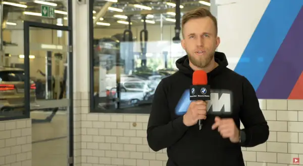 Mężczyzna z mikrofonem w serwisie BMW, informuje o akcji serwisowej. W tle widać samochody i podnośniki.
