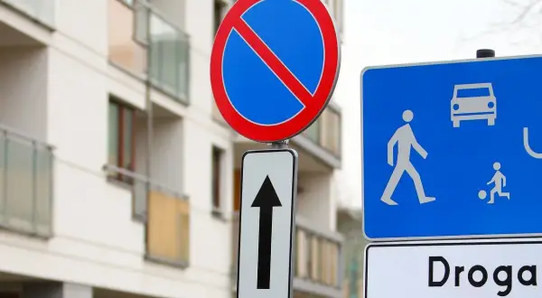 Znak zakaz postoju nad strzałką kierunkową, obok niebieskiej tablicy z piktogramami pieszych i samochodu. Budynek mieszkalny w tle.