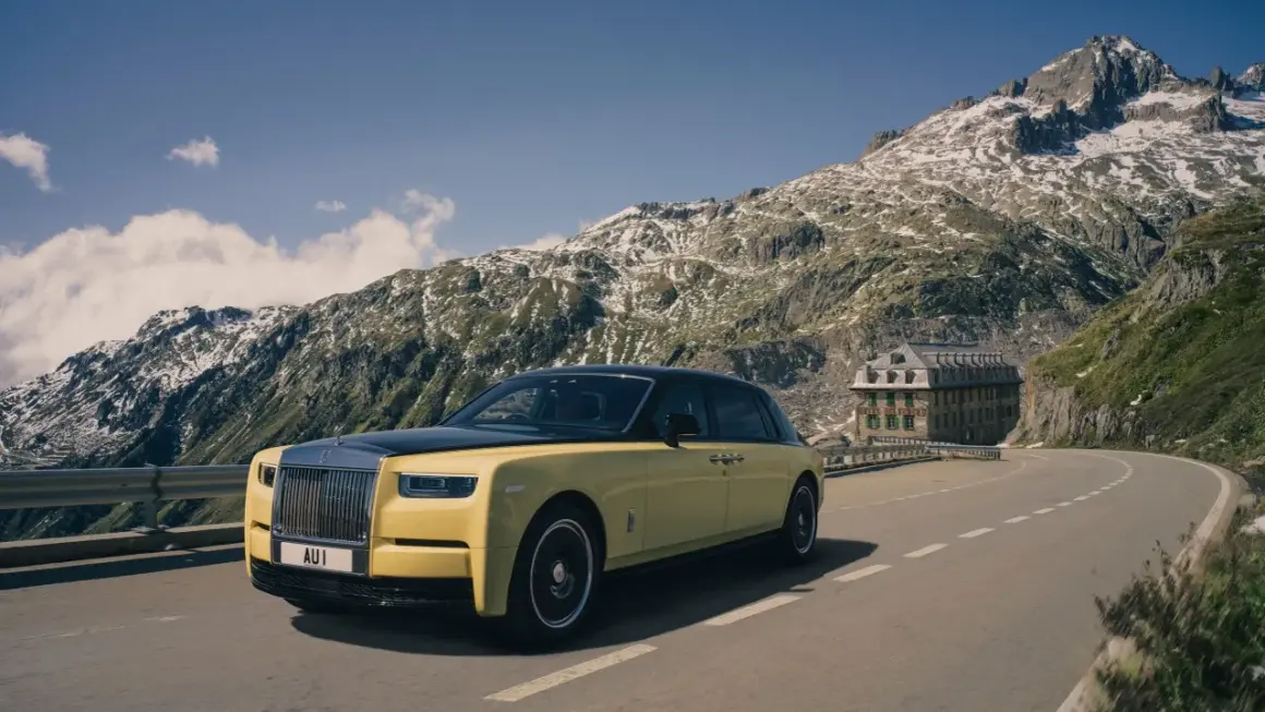 Żółty Rolls-Royce Phantom przemierza górską drogę. W tle majestatyczne szczyty i zabytkowy hotel.