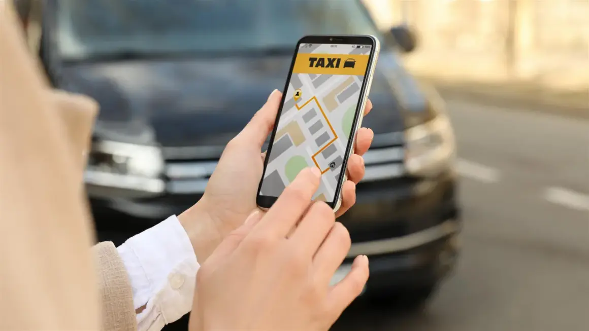 Dłoń trzyma smartfon z aplikacją taxi i mapą, na tle samochodu.