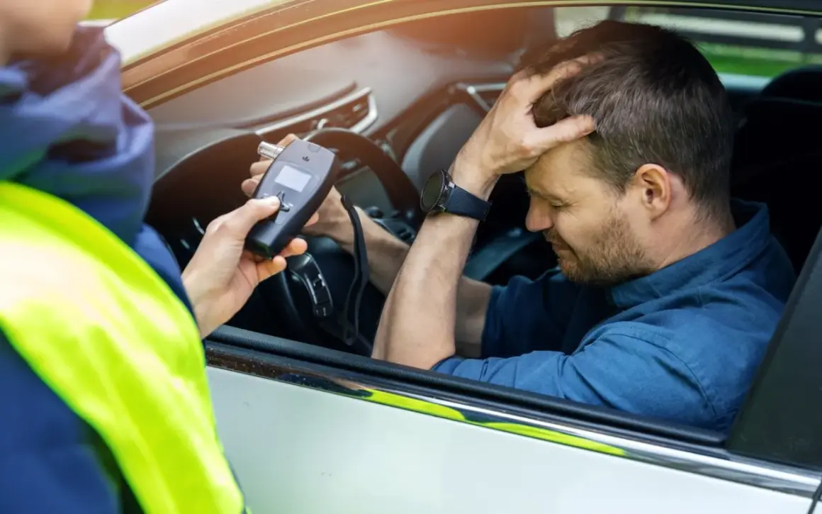 Policjant mierzy kierowcy ile promili. Mężczyzna zmartwiony trzyma się za głowę.