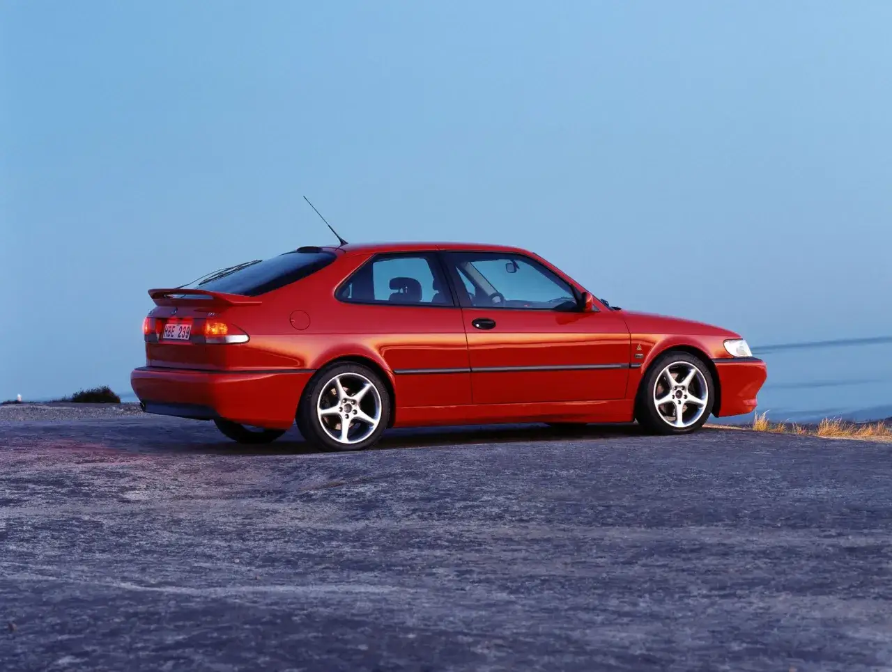 Czerwony Saab 9-3, kiedyś część koncernu General Motors, zaparkowany na kamienistym podłożu przy błękitnym niebie.