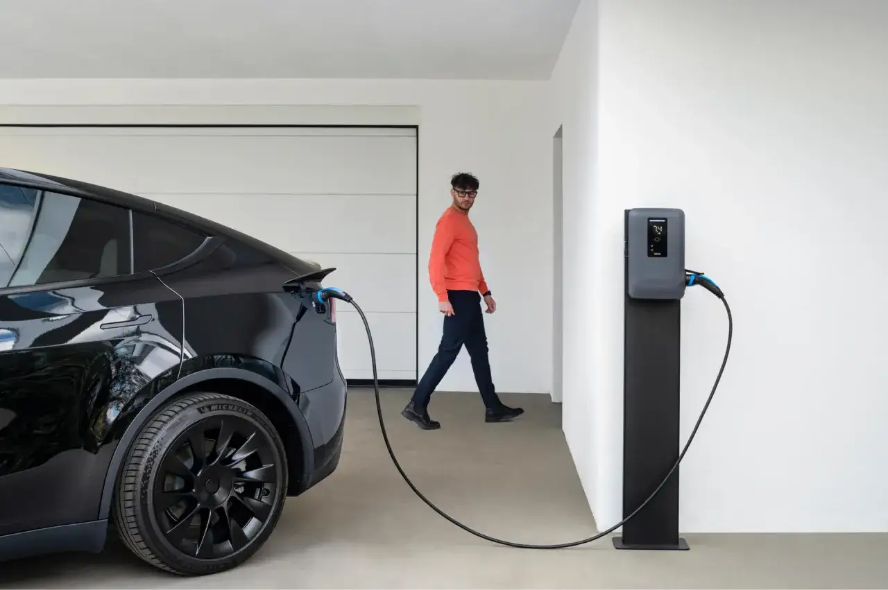 Mężczyzna w pomarańczowej bluzie obserwuje ładowanie samochodu elektrycznego w garażu.