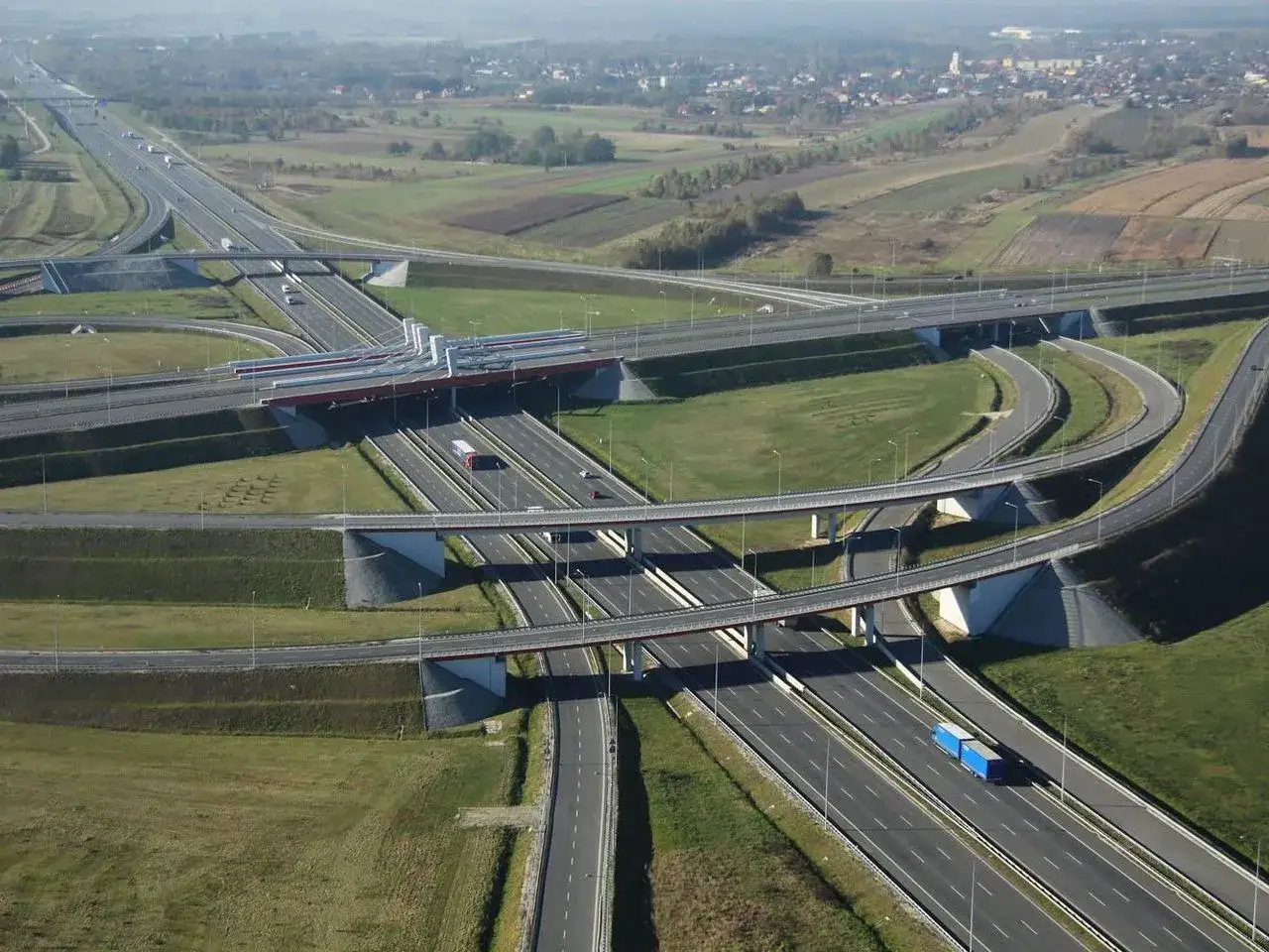 Rozległy węzeł drogowy, nowoczesna obwodnica z wieloma estakadami i zjazdami, otoczona polami i miasteczkiem w oddali.
