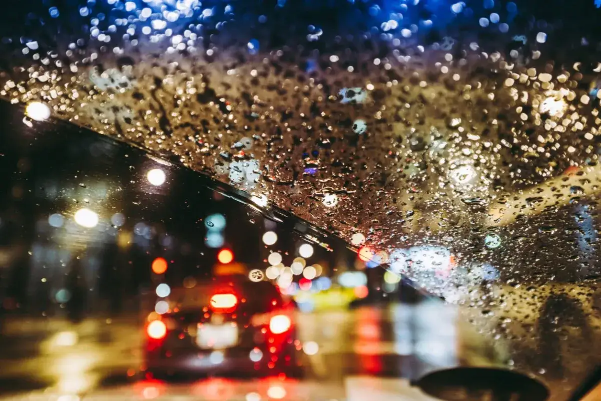 Krople deszczu na szybie rozmywają światła mijania i czerwone światła samochodów w nocnej, deszczowej podróży.