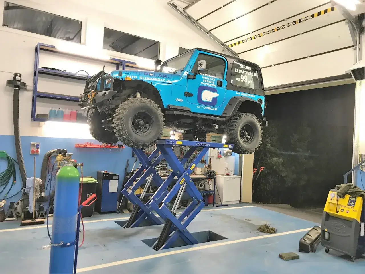 Niebieski Jeep na podnośniku w warsztacie. Na drzwiach widnieje napis 