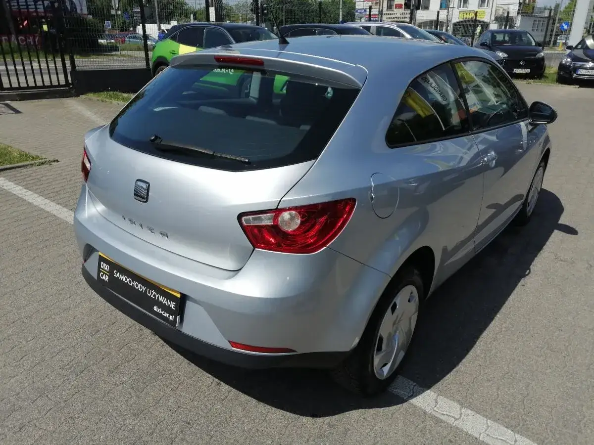 Srebrny Seat Ibiza, jeden z wielu małych samochodów na placu. W tle zielony i czarne auta.