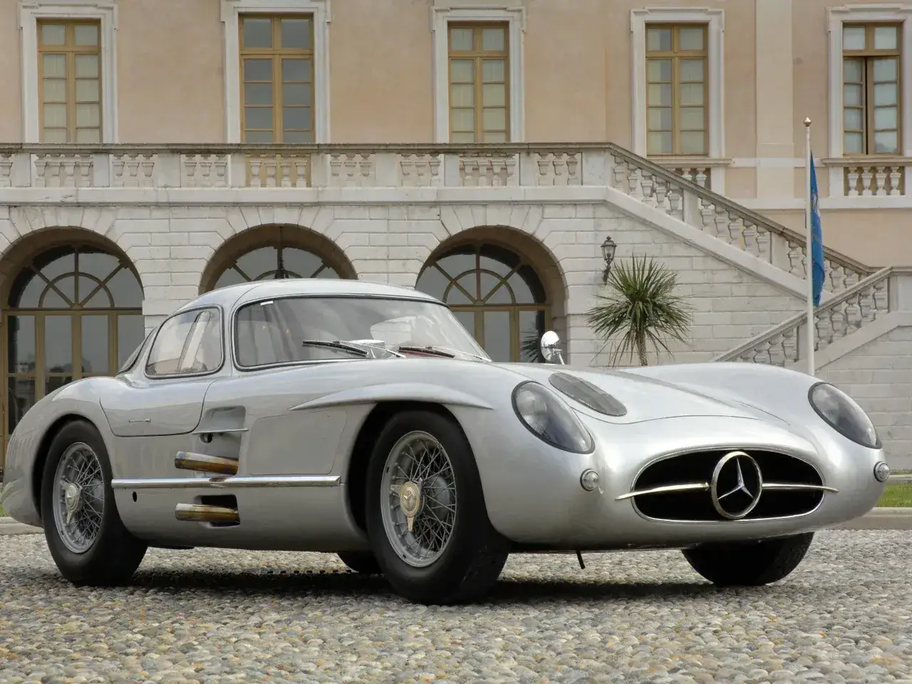Srebrny Mercedes-Benz 300 SLR Uhlenhaut Coupé, najdroższy samochód świata, przed zabytkowym pałacem.