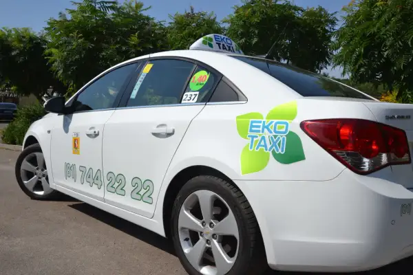 Biała taksówka EKO TAXI z zielonym logo i numerem telefonu na drzwiach.