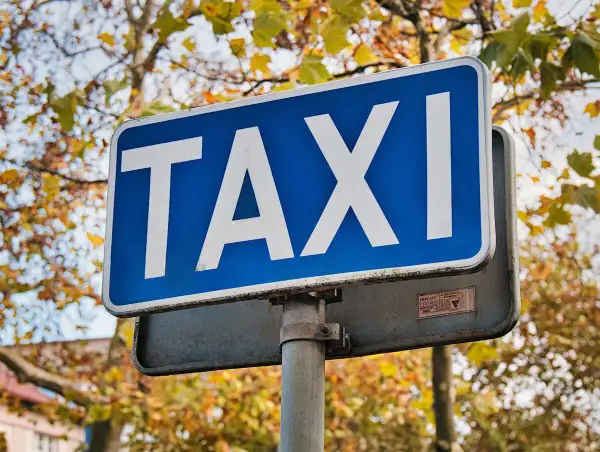 Niebieski znak TAXI z białymi literami na tle jesiennych drzew.