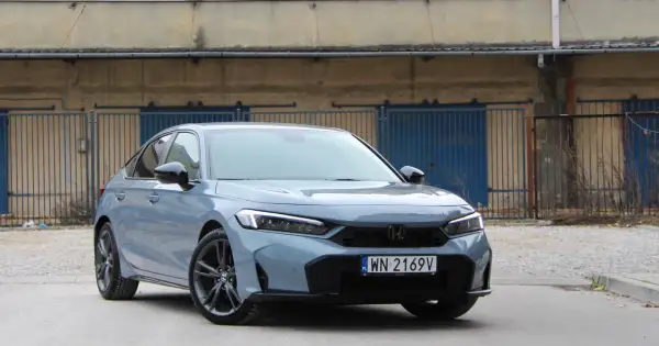 Nowy Honda Civic Sport w kolorze szarym, z czarnymi felgami, zaparkowany przed industrialnym budynkiem z niebieskimi bramami.