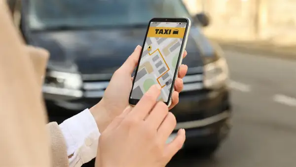 Dłoń trzyma smartfon z aplikacją taxi i mapą, na tle samochodu.