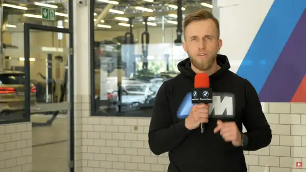 Mężczyzna z mikrofonem w serwisie BMW, informuje o akcji serwisowej. W tle widać samochody i podnośniki.