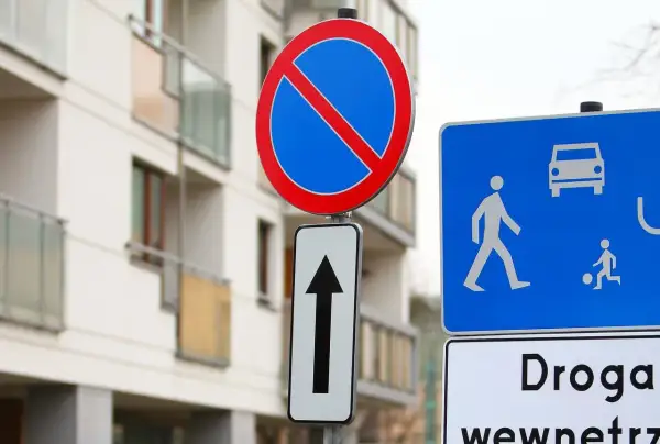 Znak zakaz postoju nad strzałką kierunkową, obok niebieskiej tablicy z piktogramami pieszych i samochodu. Budynek mieszkalny w tle.